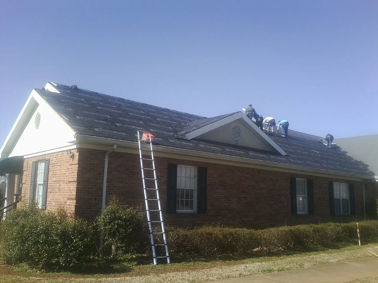 Crew tearing shingles off a brick ranch roof with a ladder set — Emergency Roof Repair work by our crew in Lonepine