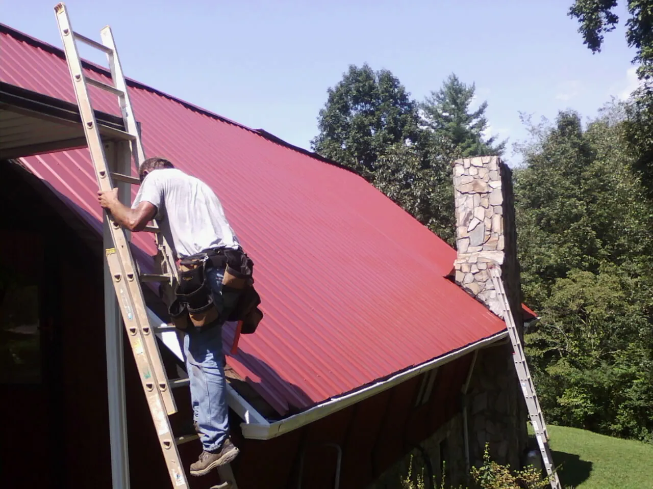 Roofer on a ladder beside a red metal roof and stone chimney — Soffit & Fascia Repair by Roofing Brothers in Lonepine